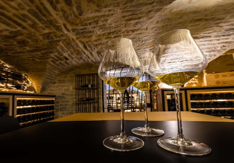 Three glasses of white wine on a table under a vaulted stone cellar roof